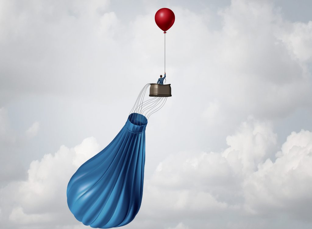 a deflated hot air balloon kept aloft by a person holding a hellium balloon