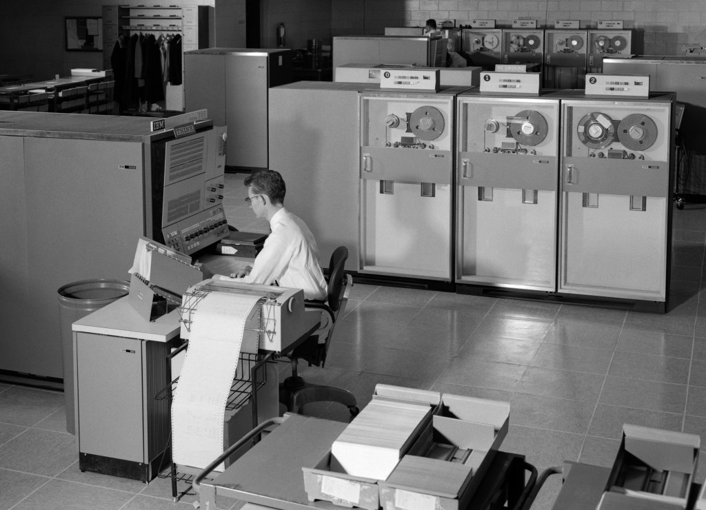 Black white photo of man sitting in front of a mainframe computer in the 1960s