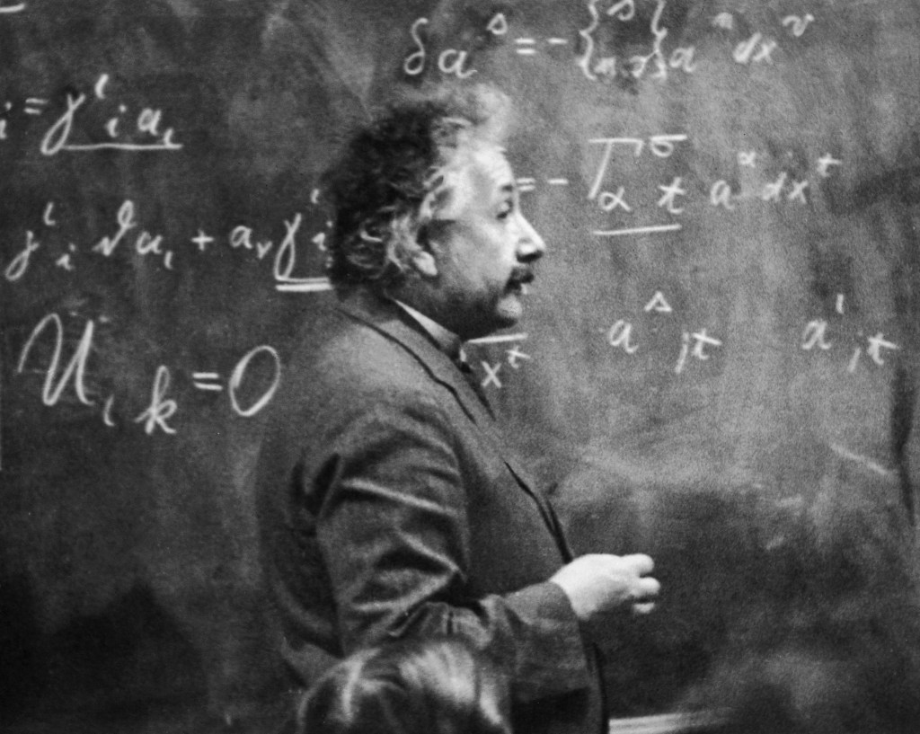 erman-born physicist Albert Einstein (1879 - 1955) standing beside a blackboard with chalk-marked mathematical calculations written across it.