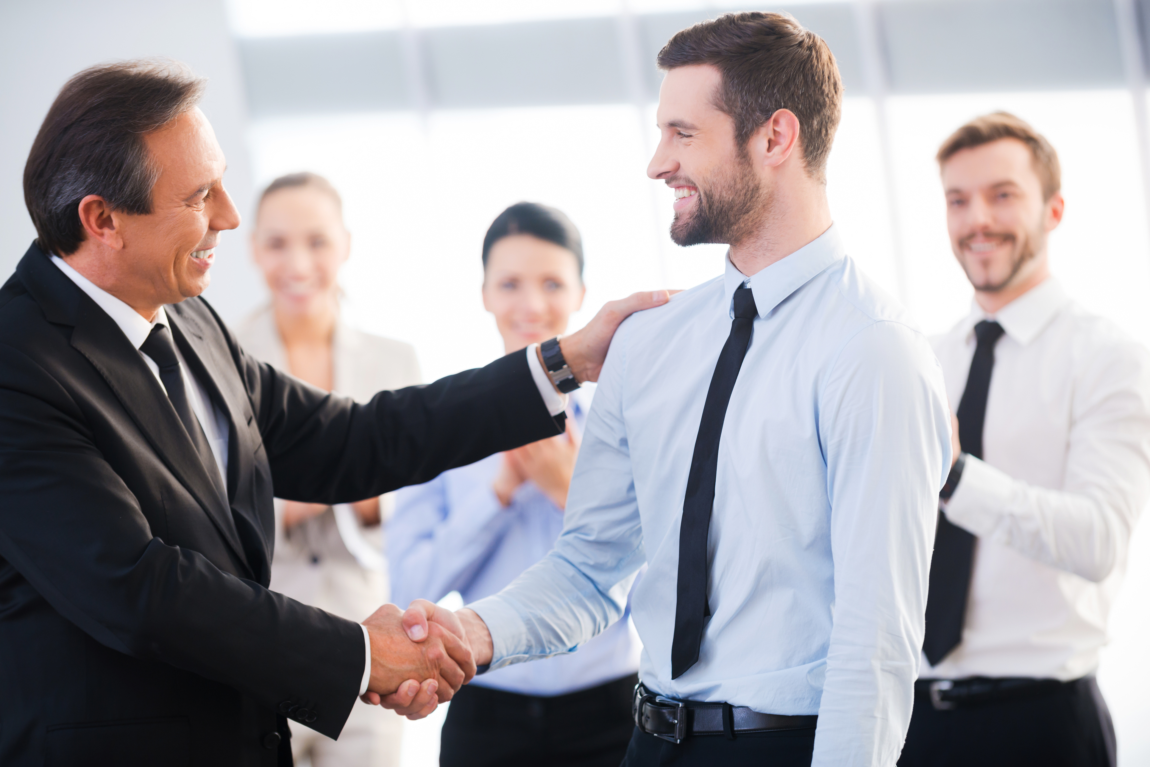 Two cheerful business men shaking hands while their colleagues applauding a...