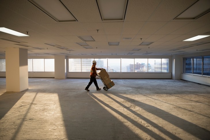 Construction worker moving cardboard boxes on handcart in sunny empty, unfinished highrise office