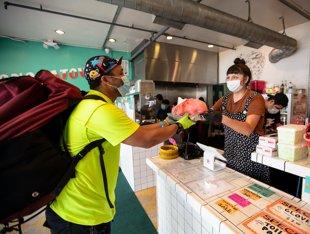 A Mexican restaurant adapts to the Covid-19 lockdown. A delivery cyclist wearing a massive backpack takes an order from behind the counter. Everyone is wearing masks and gloves.