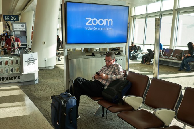 San Jose, CA, USA - Feb 13, 2020: A businessman checks his smartphone in San Jose International Airport with Zoom Video Communications advertisement on the screen in the background. Zoom provides remote conferencing services using cloud computing and has quickly emerged as one of the leading tools to keep business running and student learning.
