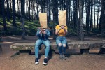 Two young woman sitting on public bench with brown bags over head using phone