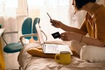 A middle-aged woman in white jeans and a yellow sweater sitting on the bed in a yoga pose in front of a laptop and a cup of coffee. Remote work at home. Online shopping in quarantine period