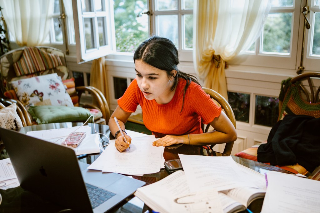 hispanic latina college student works on assignment at home. She is writing something on paper. A laptop is on her desk.