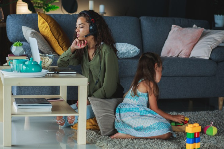 Wide shot of domestic living room where mother and daughter are sitting on the floor of the living room. Girl plays with plastic colorful toys while woman uses headphones to attend a video conference.
