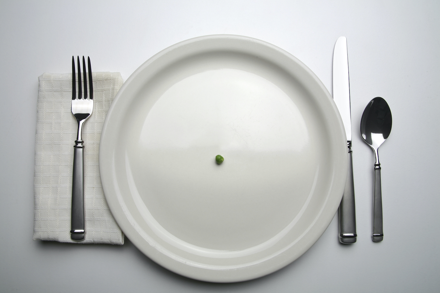 Suggesting scarcity, a single green pea rests in the middle of a dinner plate surrounded by tableware.