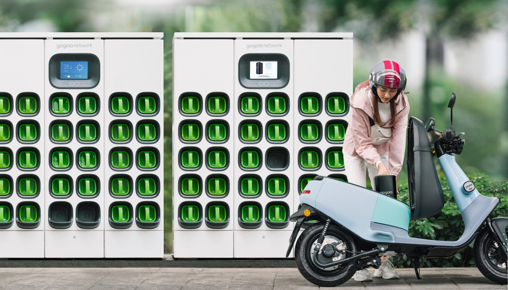 A photograph of a woman standing next to a scooter in front of a Gogoro battery swapping station