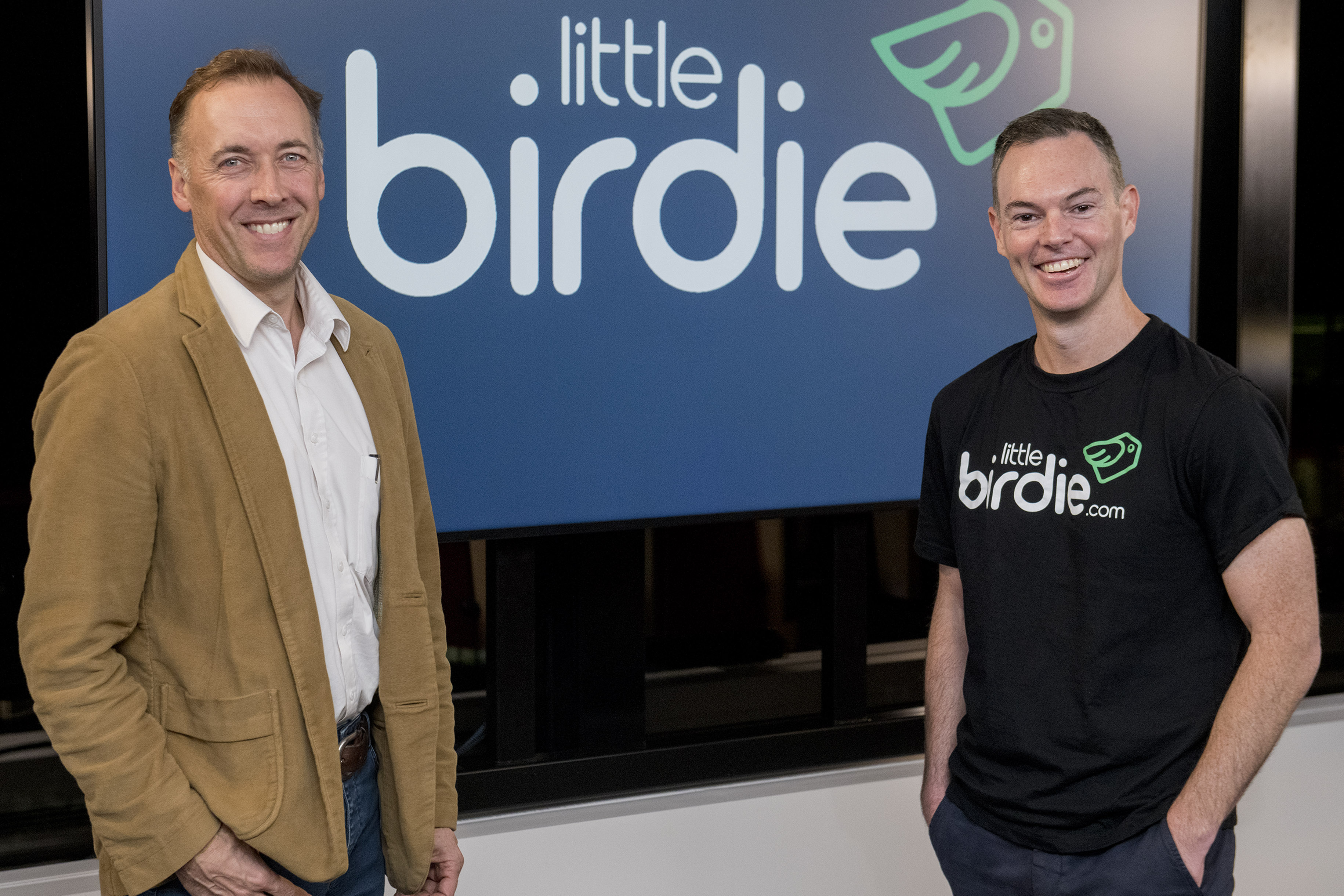 A photo of (left) Commonwealth Bank group executive Angus Sullivan and (right) Jon Beros, co-founder and CEO of Little Birdie, standing in front of Little Birdie’s logo