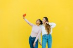 A young latinx girl and a brunette caucasian girl taking a selfie and posing together in front of a yellow background.