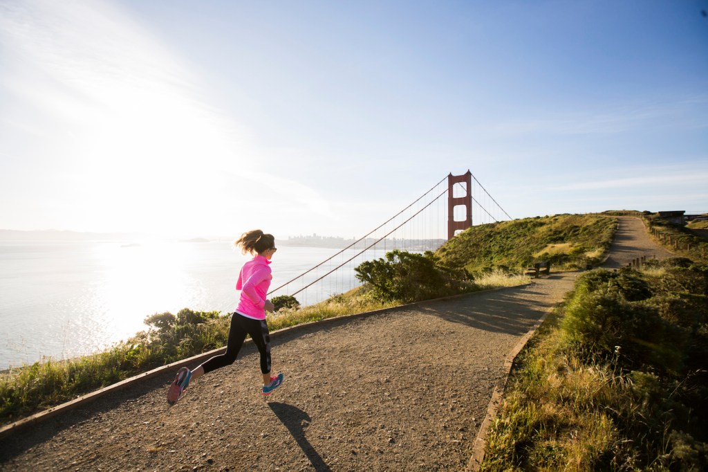 A woman running along the coast with the golden gate bridge in the distance.