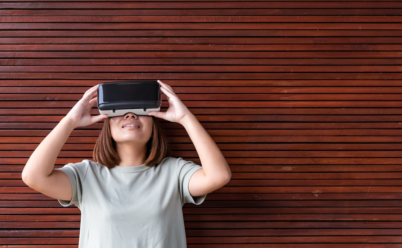Young asian woman using virtual reality simulator while standing in living room and wooden material wall background
