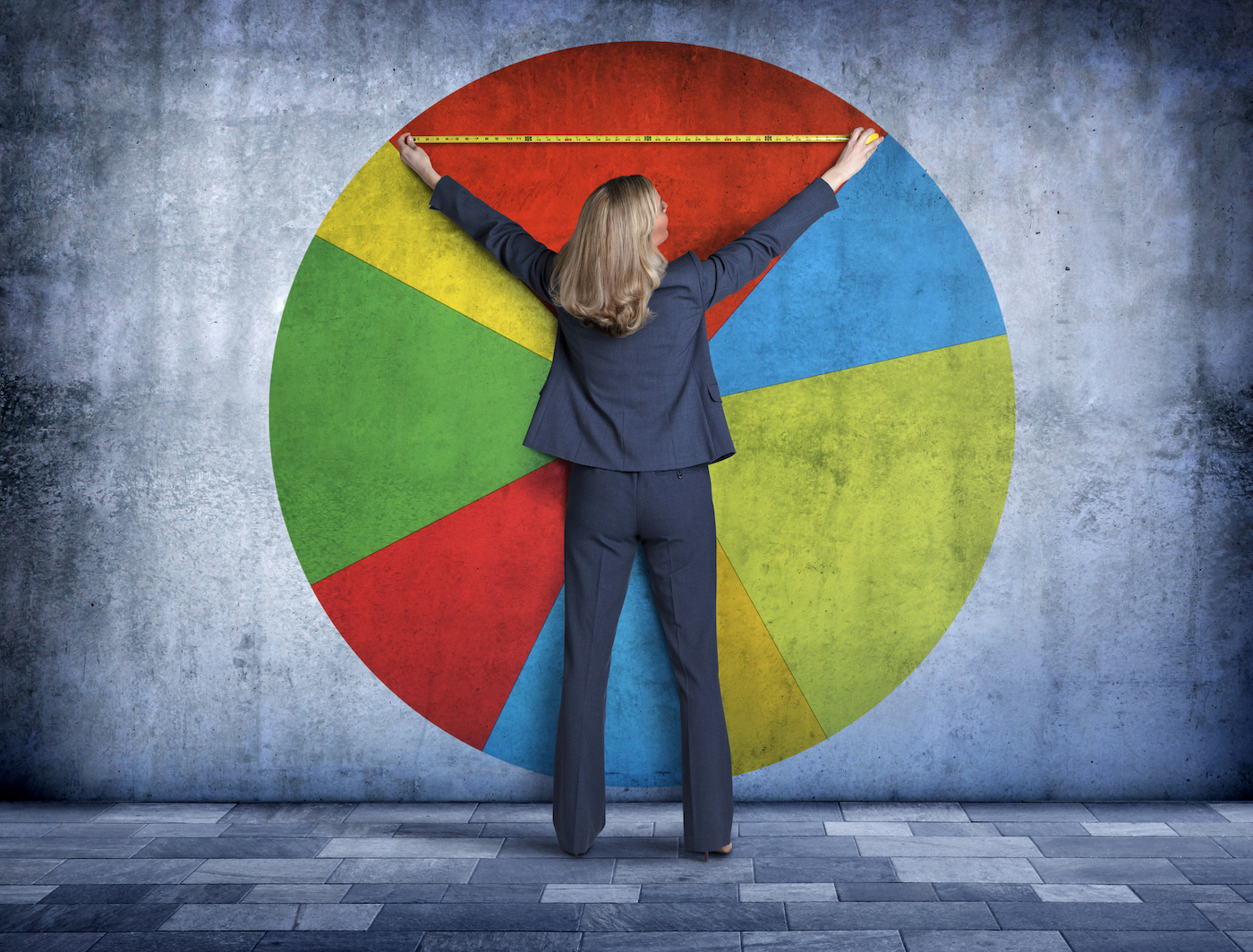 A businesswoman, using a tape measure, reaches up to measure her piece of the pie on a large pie chart that is projected onto a concrete wall.