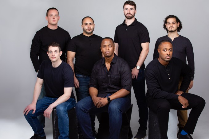 Seven people sit wearing all black staring at the camera. Founder Benjamin Harvey is in the middle.