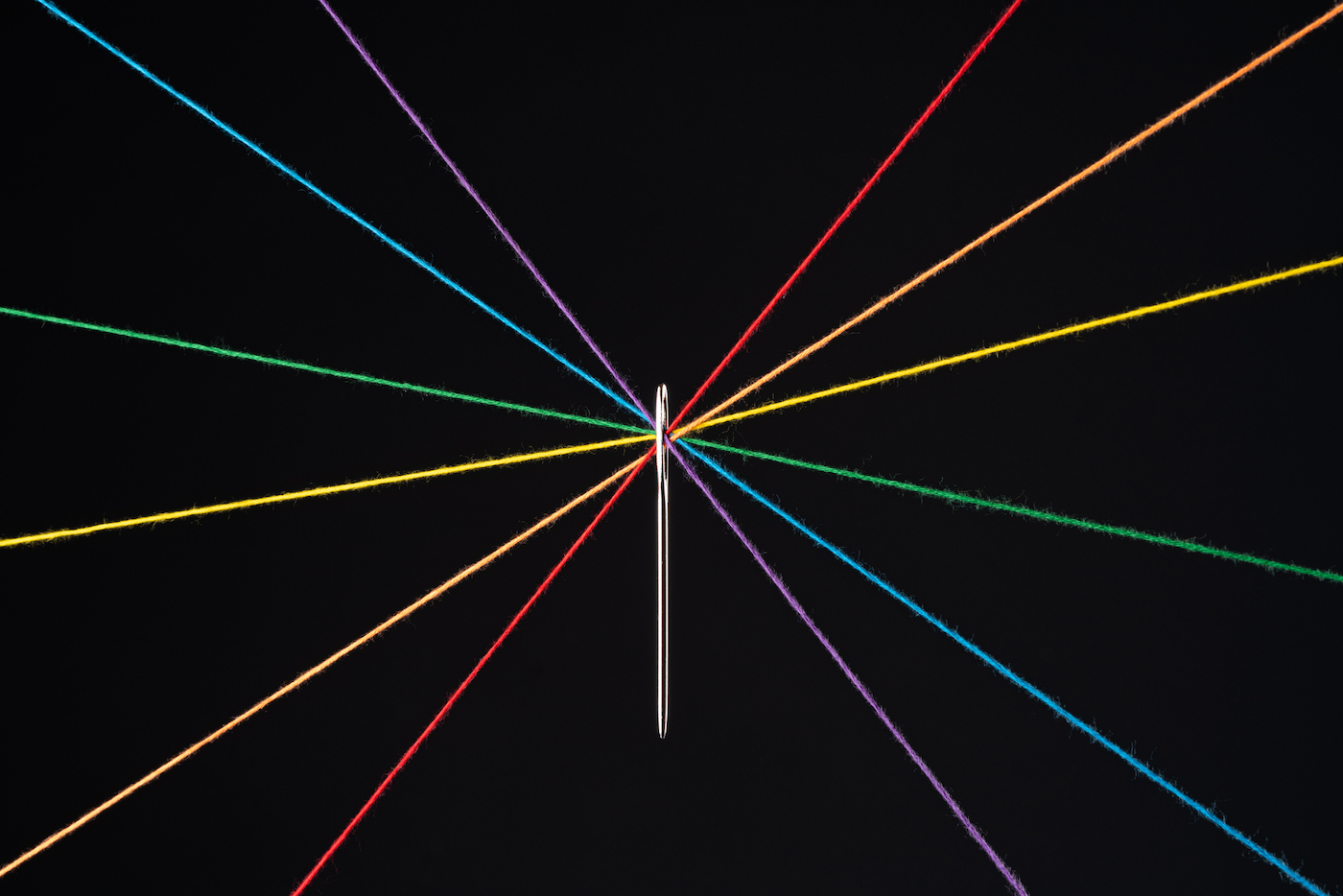 Rainbow colored threads through the eye of the needle on a black background.