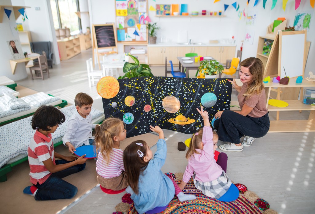 High angle view of preschool teacher showing poster with planets to children indoors in nursery, used in a post about Winnie's job marketplace