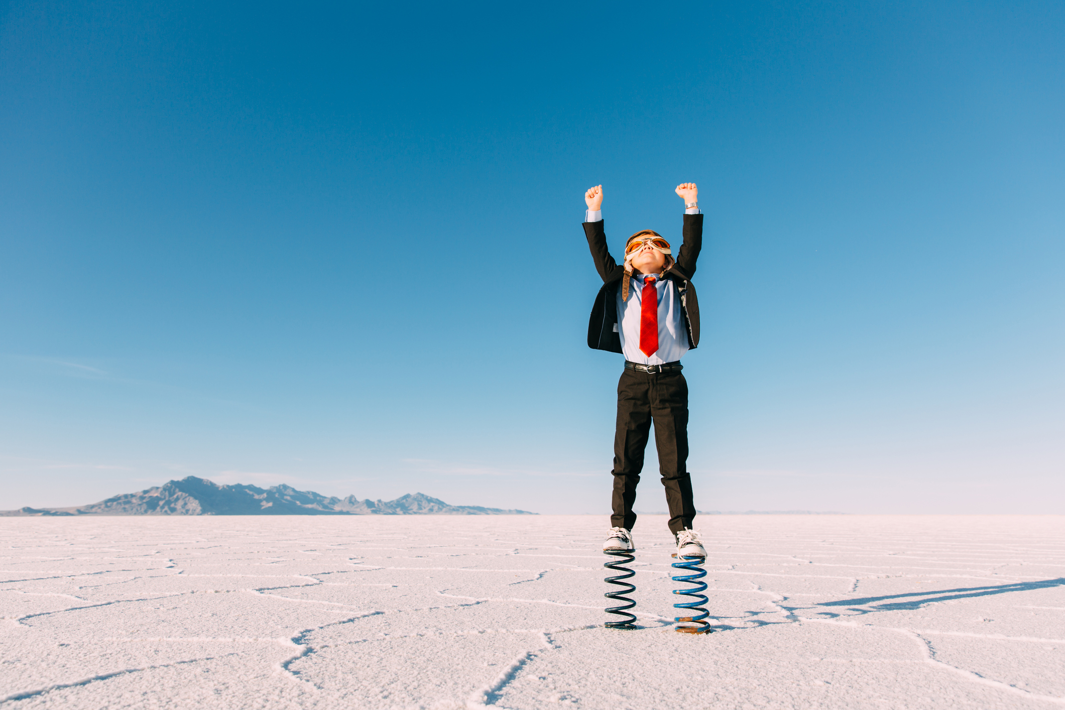 A young business boy dressed in a business suit, flight cap and goggles stands on springs in the Utah desert.  growing up enterprise for saas startups