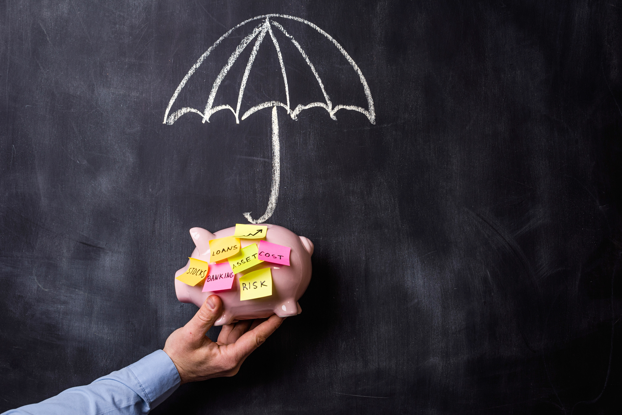 Hand holding Piggy bank with post it notes in front of blackboard showing a hand drawn umbrella. insurtech