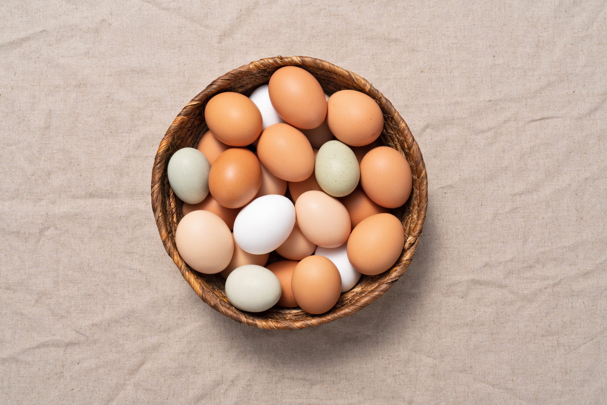 Multicolored chicken eggs in a basket on a beige colored linen tablecloth.