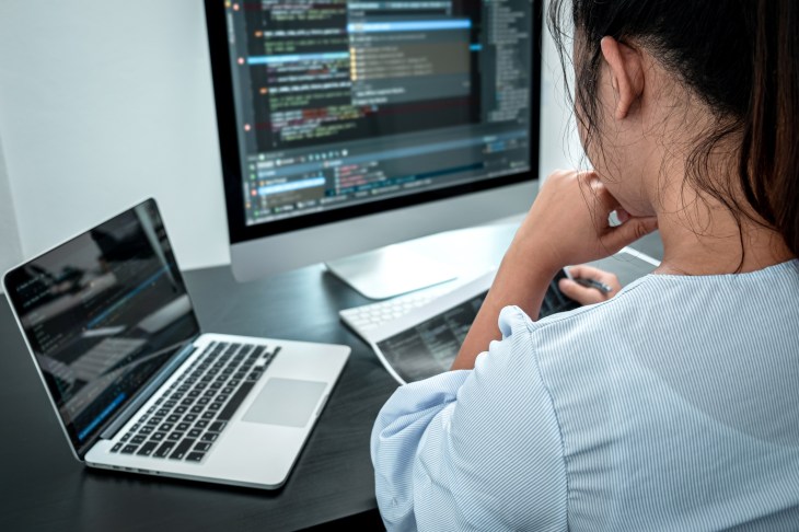 Woman coding an application on a laptop with large dual monitor next to it.