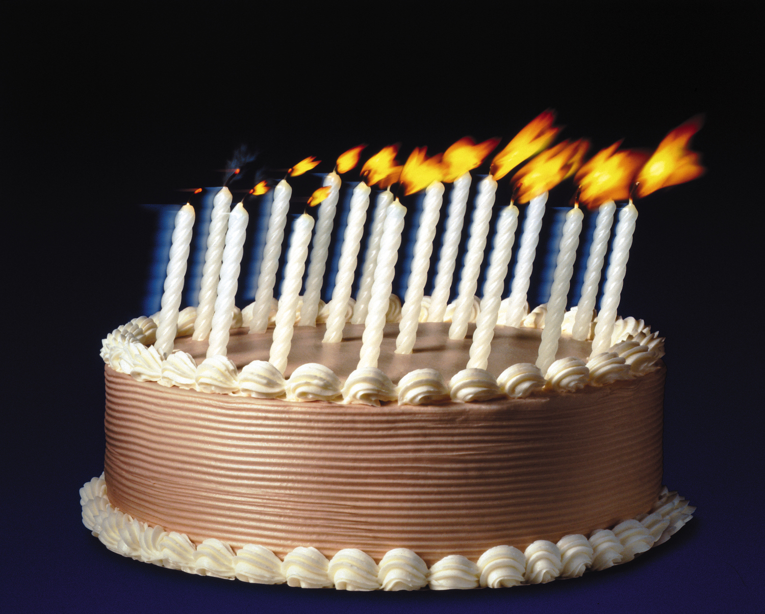 A frosted cake with candles that are being blown upon as if someone just out of frame were making a wish