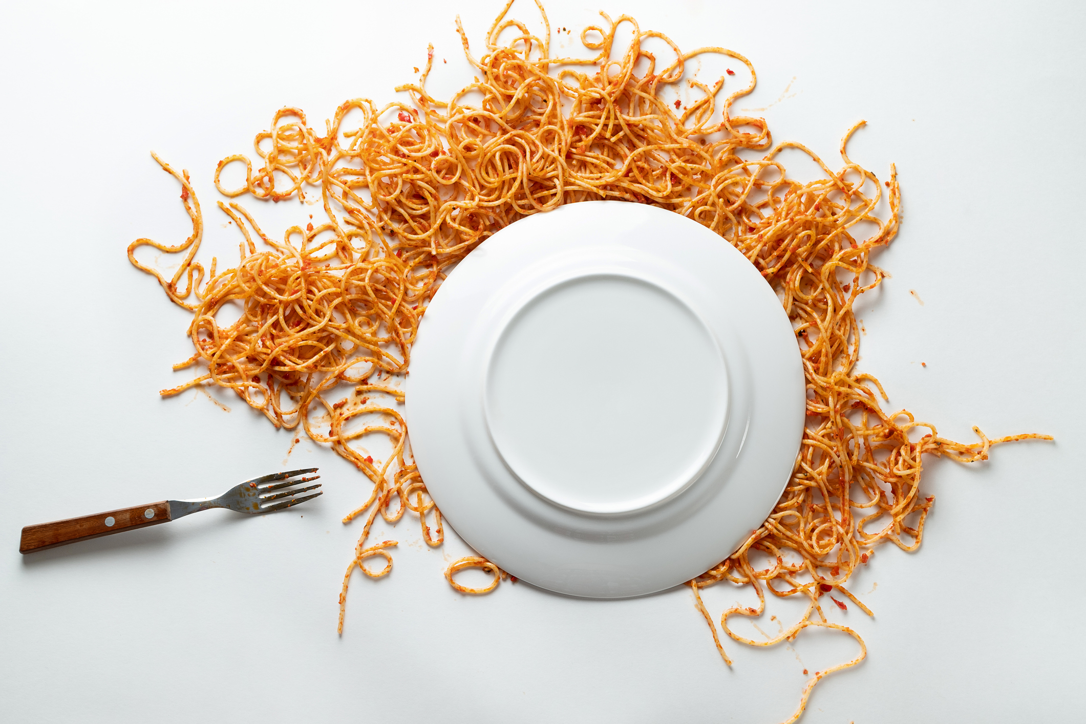 옆에 포크가 있는 스파게티의 타락한 흰색 세라믹 접시.  토마토 소스의 파스타 볼로네제는 흰색 배경이나 테이블에 흩어져 있습니다.  채식과 완전 채식의 개념.  음식 배경.  텍스트 공간의 사본.
