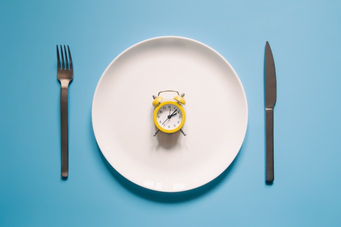 Alarm clock on a white plate with a knife and fork on blue background. Intermittent fasting, Ketogenic dieting, weight loss, meal plan, and healthy food concept.