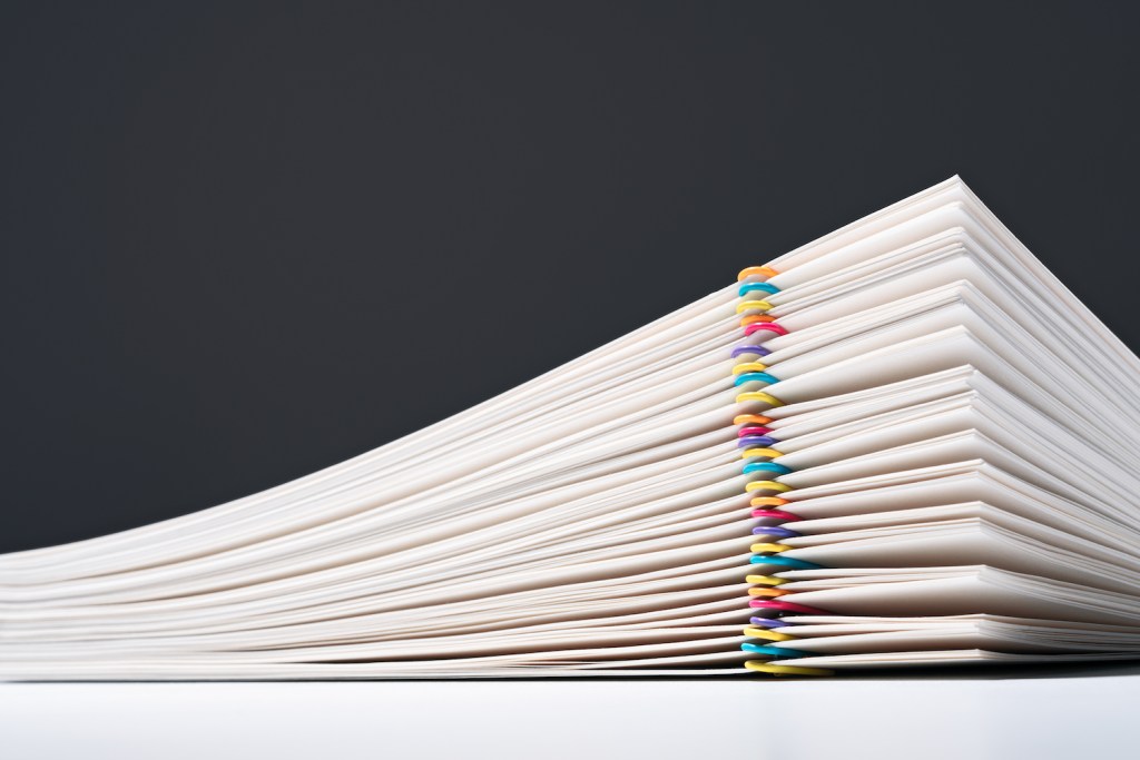 Abundance of Stacked Paper Files With Multi Colored Paper Clips Side View Against Gray Background.