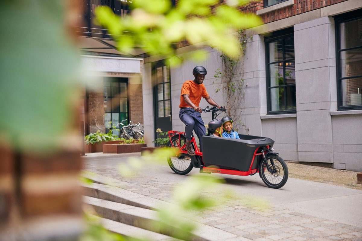 Father rides front-loading cargo e-bike with two small children inside.