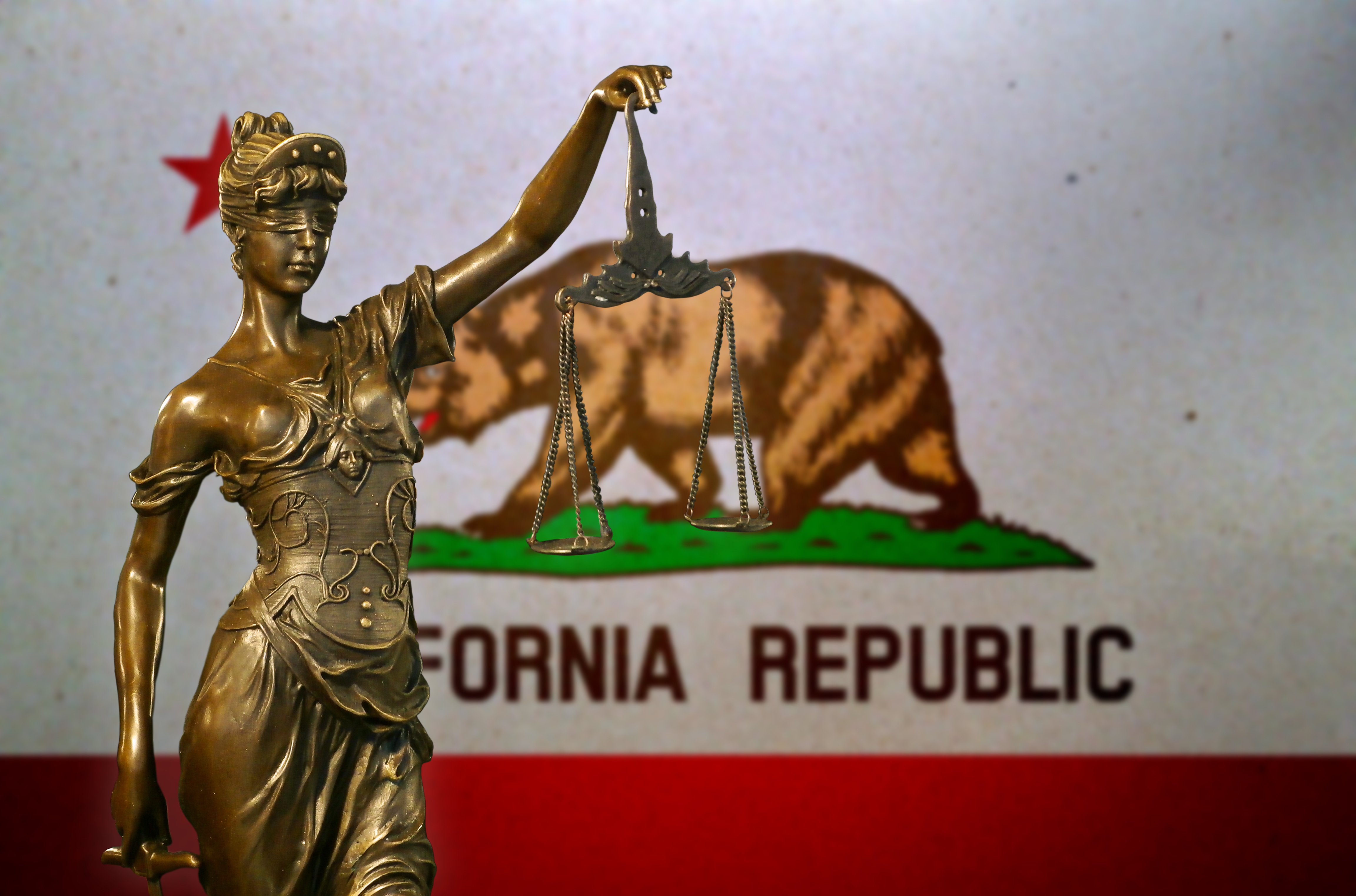 Close-up of a small bronze statuette of Lady Justice before a flag of California.