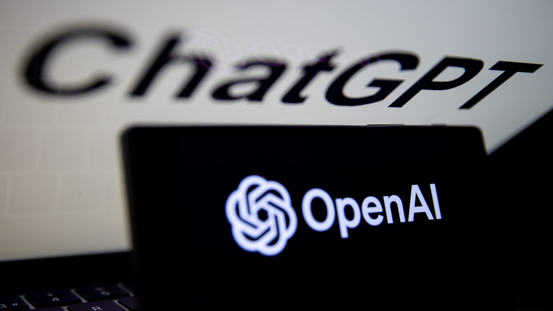 The OpenAI logo is displayed on a mobile phone screen in front of a computer screen with ChatGPT's logo