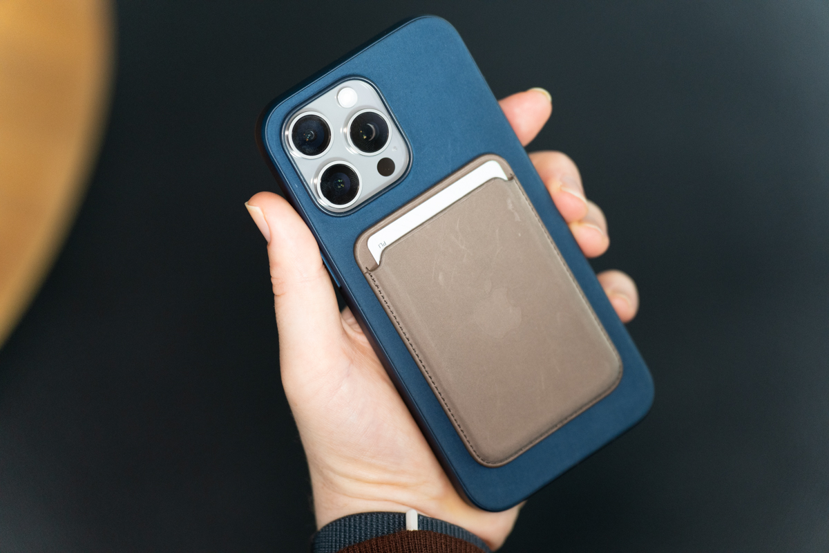 iPhone 15 Pro Max in a blue fabric case, with a MagSafe wallet accessory, being held and showing the back of the phone