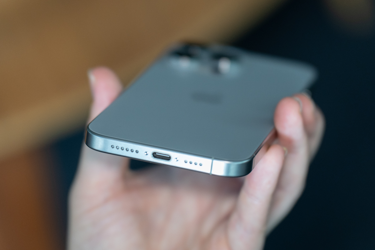 iPhone 15 Pro Max in natural titanium, being held, showing the bottom of the phone and USB-C port