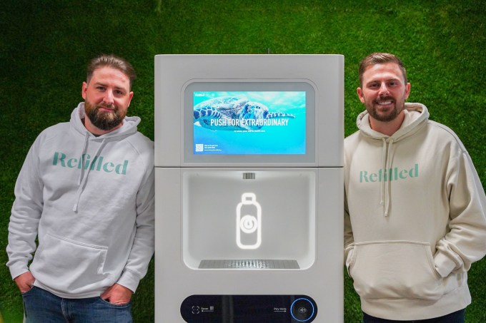 A photo of two men standing with a Refiller drink dispenser between them, against a green background