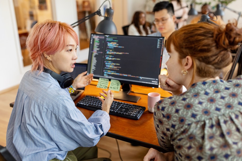 Female software developers discuss over the computer while sitting at a desk in the workplace. Creative businesswomen discuss the new coding program in the office.