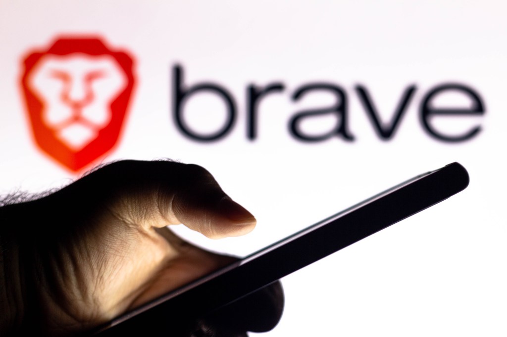 In this photo illustration, a hand of a person holds a smartphone with a Brave Browser logo displayed in the background.